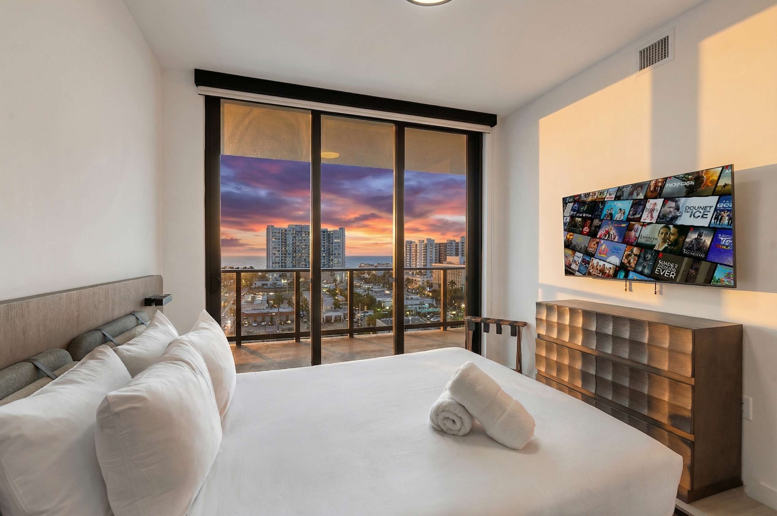 A modern hotel room with a large bed, a city view, a wall-mounted TV, and a wooden headboard.