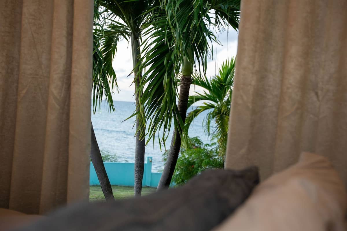 A room with a view of the ocean through curtains, a bed, and a window.