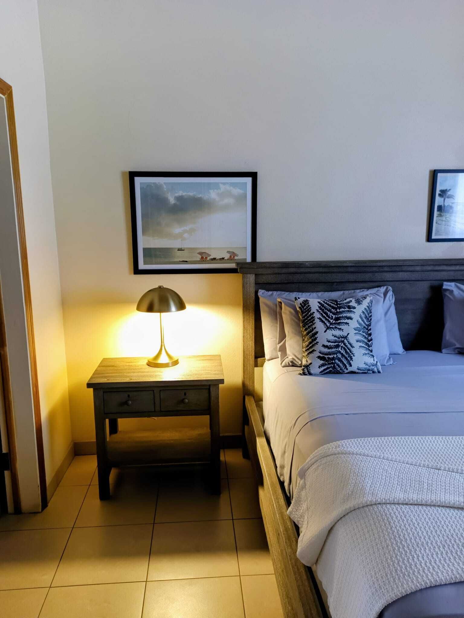 Een keurig opgemaakt slaapkamer met een bed, een nachtkastje met een lamp, en twee ingelijste foto's aan de muur.