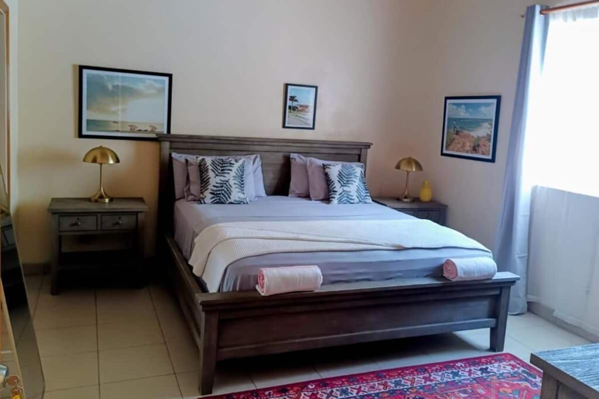 A bedroom with a wooden bed, two bedside tables with lamps, a large window with curtains, and two framed pictures on the wall.