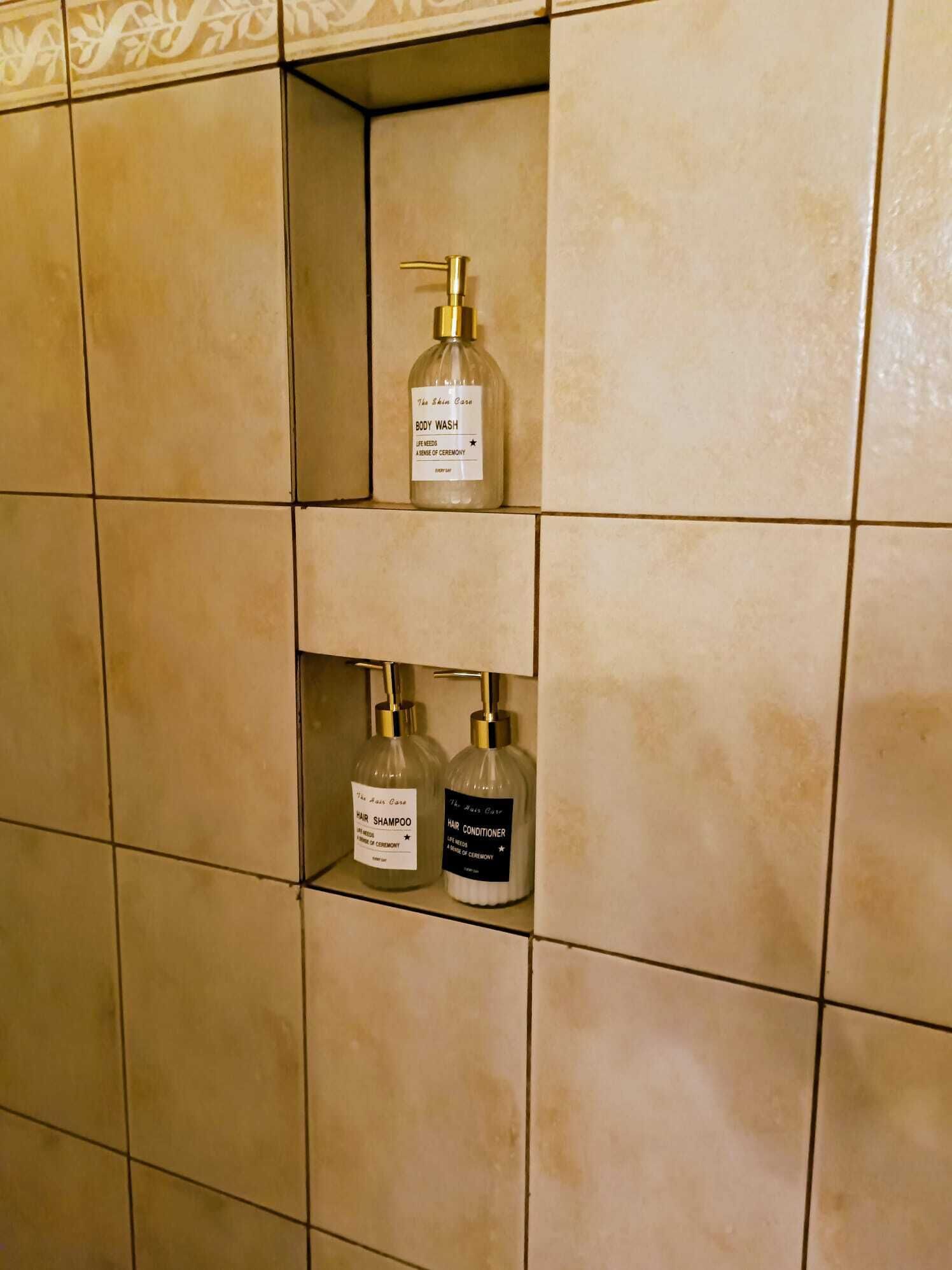 A bathroom with beige tiles and three bottles of The Hair Care brand products: body wash, hair shampoo, and hair conditioner.