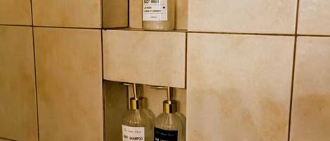 A bathroom with beige tiles and three bottles of The Hair Care brand products: body wash, hair shampoo, and hair conditioner.