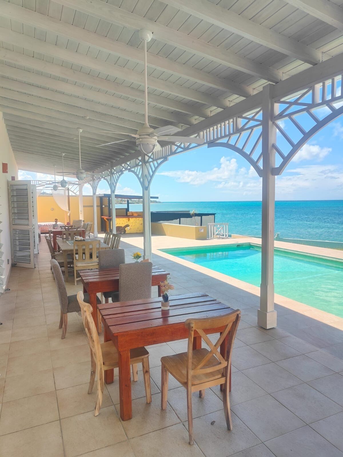 A covered outdoor area with wooden tables and chairs, a pool, and a view of the ocean. - Turtle Cove House by Le Haus (St. John's)