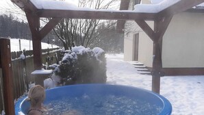 A wooden hot tub in a backyard with snow on the ground and a wooden pergola overhead.