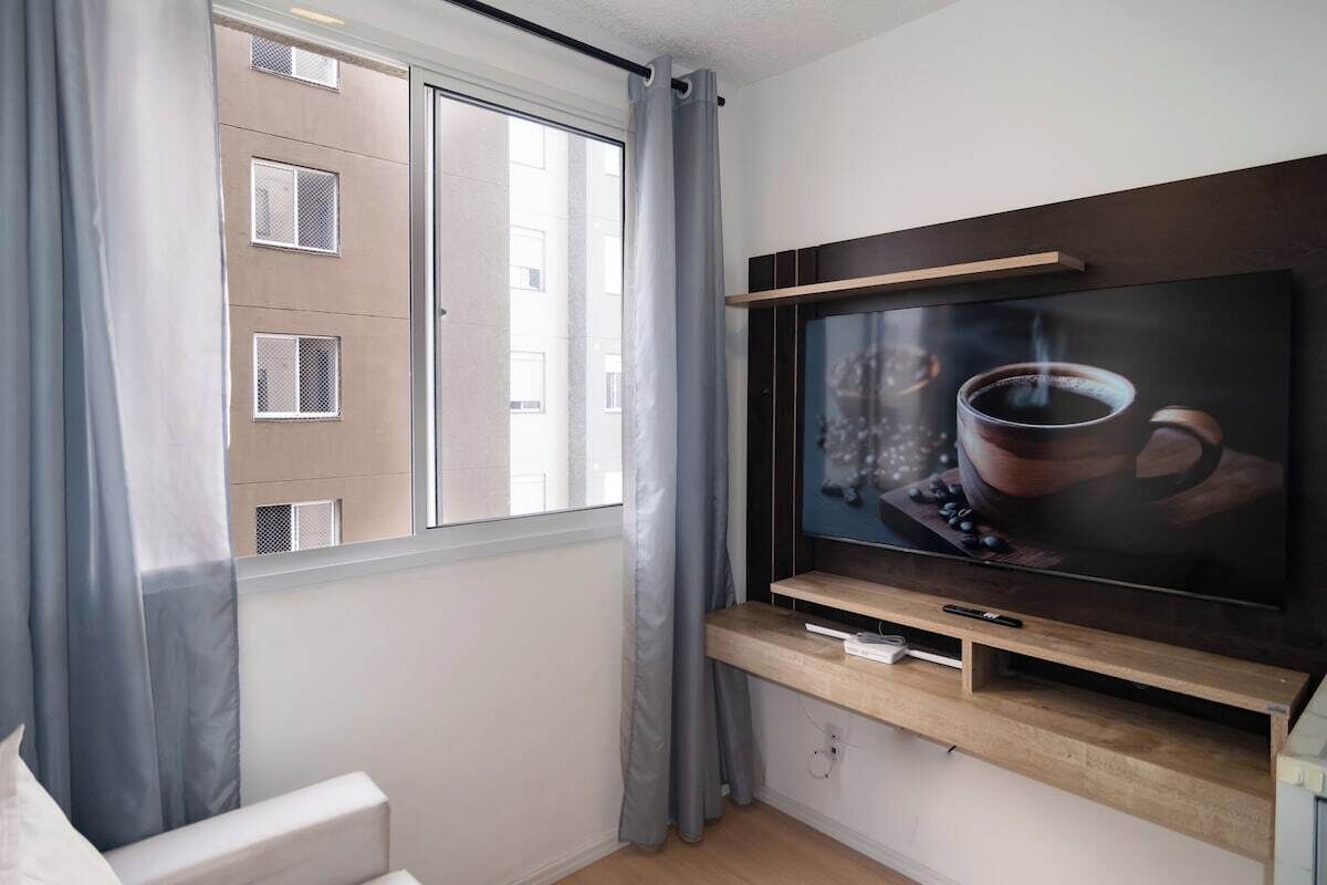 Uma sala de estar moderna com uma televisão de tela plana grande presa em uma prateleira de madeira, uma janela com cortinas cinzas e vista para um edifício.