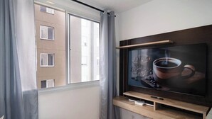 A modern living room with a large flat-screen TV mounted on a wooden shelf, a window with grey curtains, and a view of a building outside. - Cozy Apartment for 2 (between CGH and Interlagos) (São Paulo)