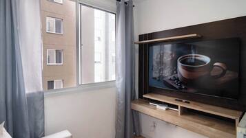 Uma sala de estar moderna com uma televisão de tela plana grande presa em uma prateleira de madeira, uma janela com cortinas cinzas e vista para um edifício.