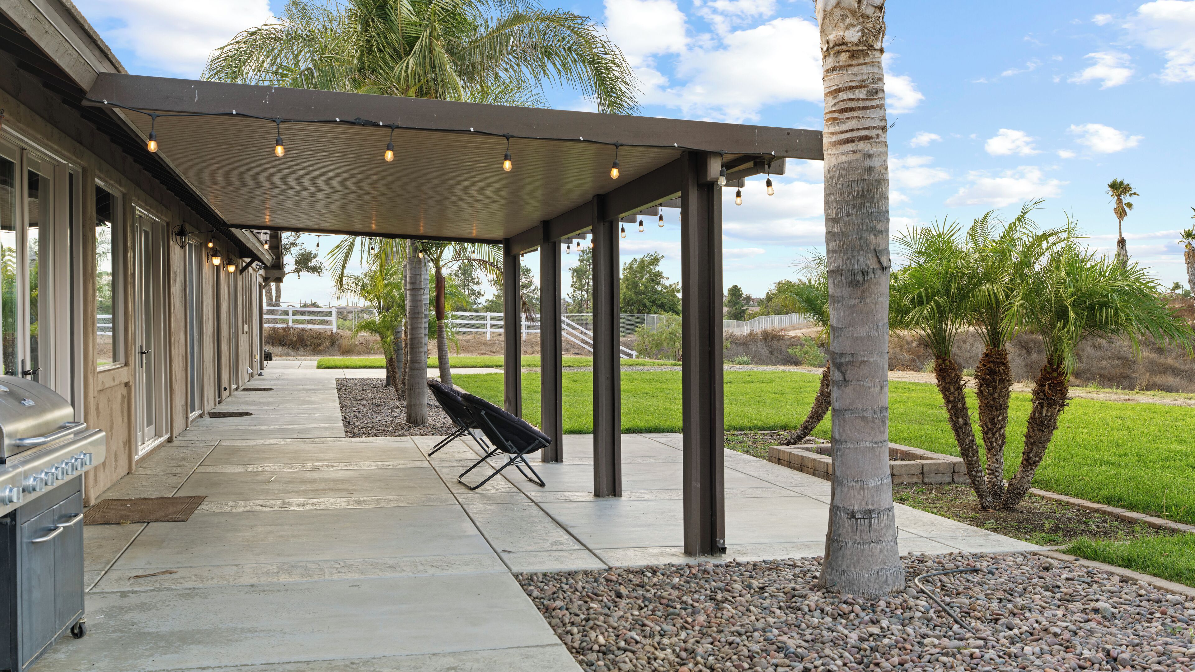 A covered patio area with a grill, outdoor seating, and palm trees.