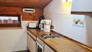 A compact kitchen with wooden ceiling, white cabinets, a built-in oven, a coffee maker, and a window with blinds. - Cozy vacation apartment in Reit im Winkl - perfect for your vacation (Reit im Winkl)