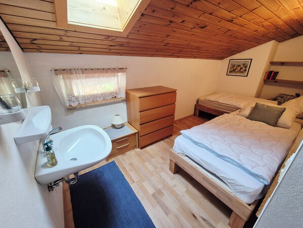 Quarto com teto de madeira, cama, mesa de cabeceira com luminária, cômoda e janela com cortina.