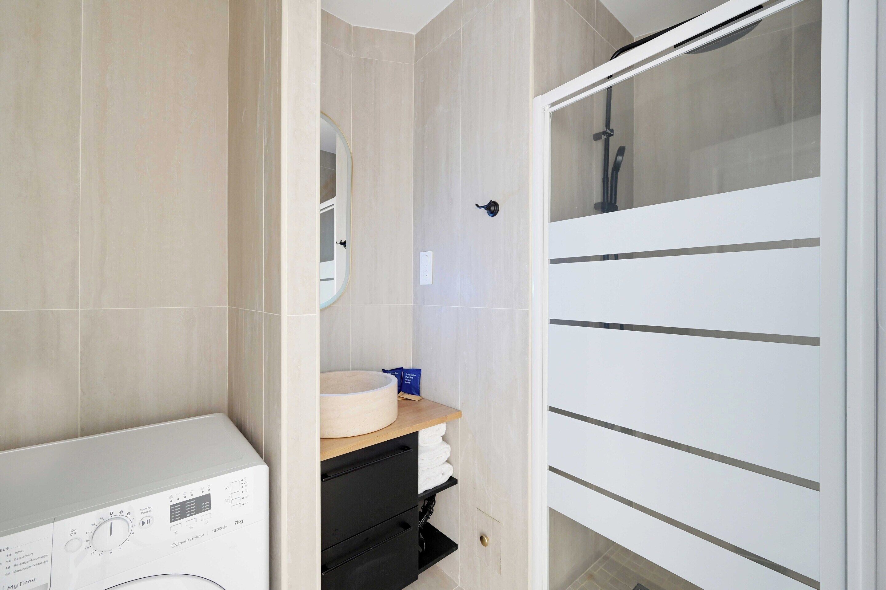 Une salle de bain moderne dotée d’un lave-linge intégré, d’un meuble-lavabo en bois et d’une cabine de douche en verre.