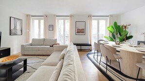 A modern living room with a sofa, dining table, and chairs. There are large windows with curtains, a plant, and artwork on the walls. - Superb apt A/C - 2br/6p - St Germain-des-pres (Paris)