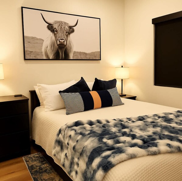 A hotel room with a bed, two bedside tables, a lamp, a framed picture of a bull, and a large window.