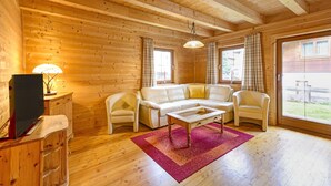 A cozy log cabin interior with a wooden floor, a sofa set, a television, and a large window with curtains. - Holiday Village Wildschonau Nature Escape Chalet (Wildschönau)