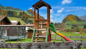 A playground with a slide and swings, surrounded by a wooden fence, set against a backdrop of green hills and a clear sky. - Holiday Village Wildschonau Nature Escape Chalet (Wildschönau)
