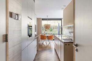 Una cocina moderna con piso de madera en patrón de espiga, muebles blancos y un comedor con dos sillas anaranjadas.