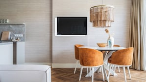 A modern dining area with a wooden herringbone floor, a white dining table, and orange upholstered chairs. A flat-screen TV is mounted on the wall above the table. - Inviting Apartment With a Garden (Sint Maartensvlotbrug)