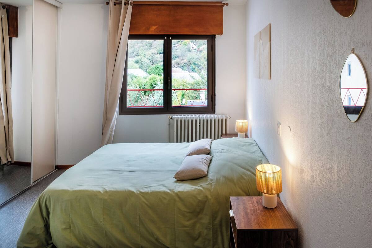 A bedroom with a bed, a window with a view of greenery, a bedside table with a lamp, and a mirror on the wall.