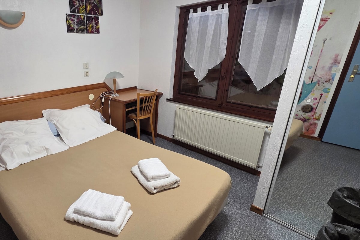 A hotel room with a bed, bedside table, lamp, and a window with curtains.