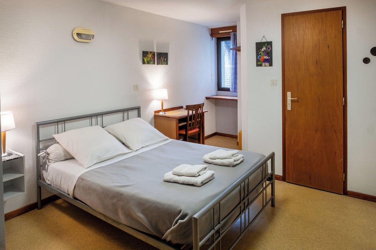 A neatly made bed with two pillows and two folded towels, a desk with a chair, a wooden door, and a small window with curtains.