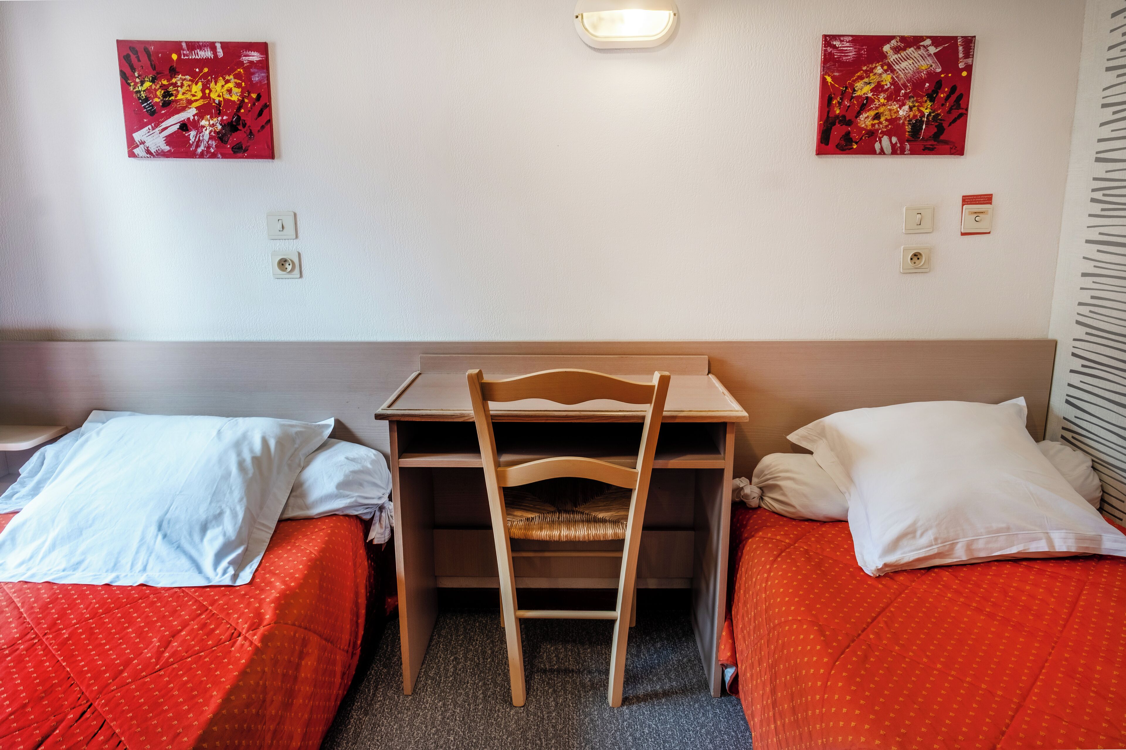A hotel room with two single beds, a wooden desk, and two paintings on the walls.