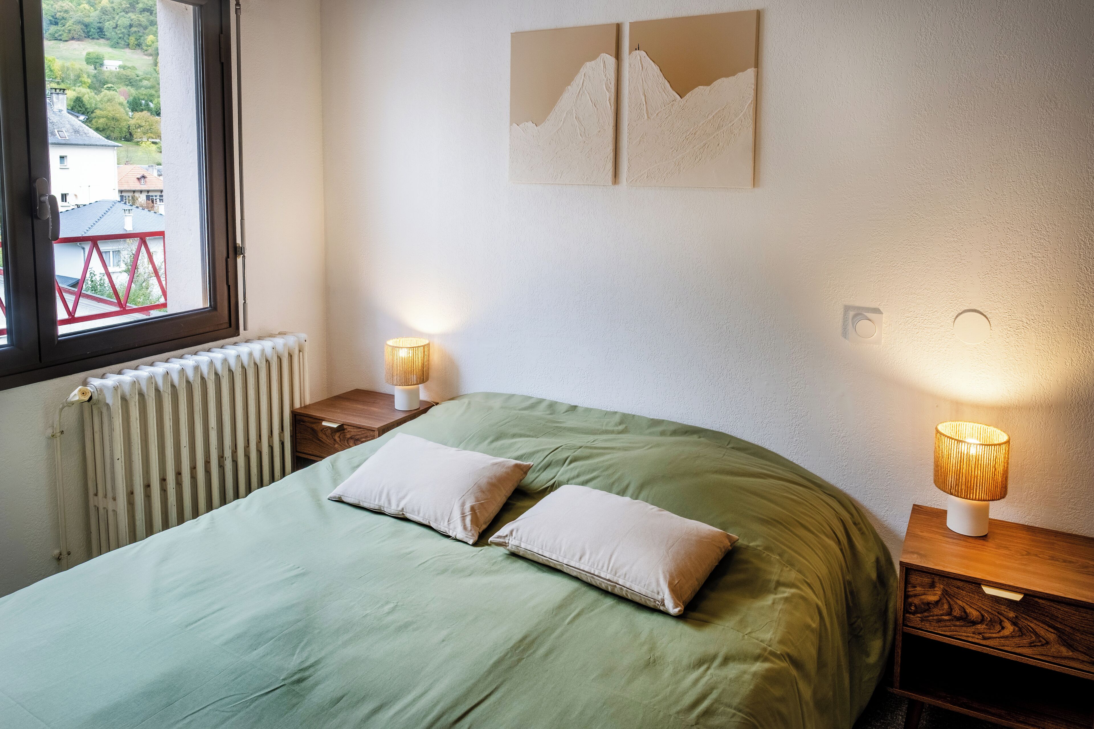 Un dormitorio con cama verde, dos almohadas, una mesita de noche de madera con lámpara y un radiador.