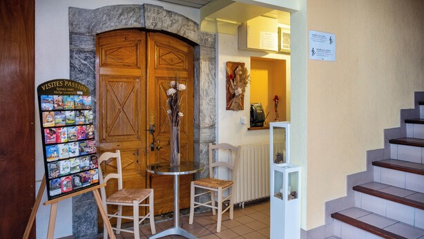 A reception area with a wooden door, a bulletin board with brochures, two chairs, a small table, and a staircase. - Hotel des Touristes (Pierrefitte-Nestalas)
