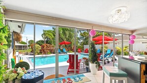 A poolside area with a glass-enclosed patio, red lounge chairs, a bar area, and a view of a swimming pool. - Waterfront w/ Htd Pool, Tiki Hut, Billards, & Bar (Boynton Beach)