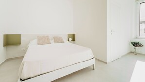 A minimalist bedroom with a white bed, two pillows, a bedside table with a lamp, and a small plant by the window. - Hieron II (Siracusa)