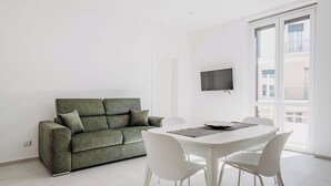 A modern living room with a green sofa, white chairs, a dining table, and a TV mounted on the wall. - Hieron II (Siracusa)