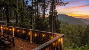 A wooden deck with string lights, overlooking a forested area with a sunset view.