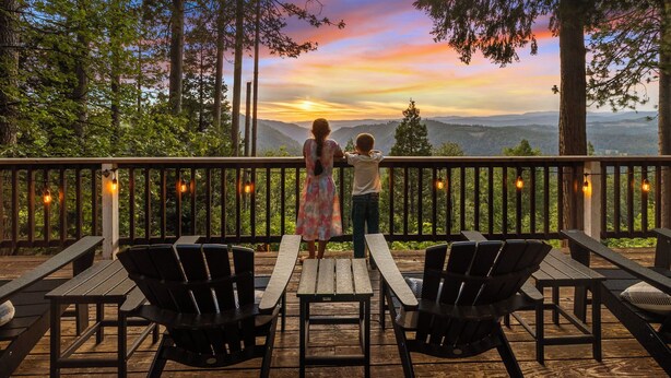 Eine Holzterrasse mit Picknicktischen und -stühlen, mit Blick auf eine bewaldete Landschaft und einen Sonnenuntergang.