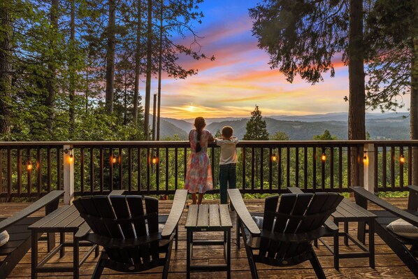 A wooden deck with picnic tables and chairs, overlooking a forested landscape with a sunset view.