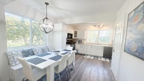 Una cocina moderna con armarios blancos, un comedor con mesa y banco blancos, y vistas al exterior a través de amplios ventanales.