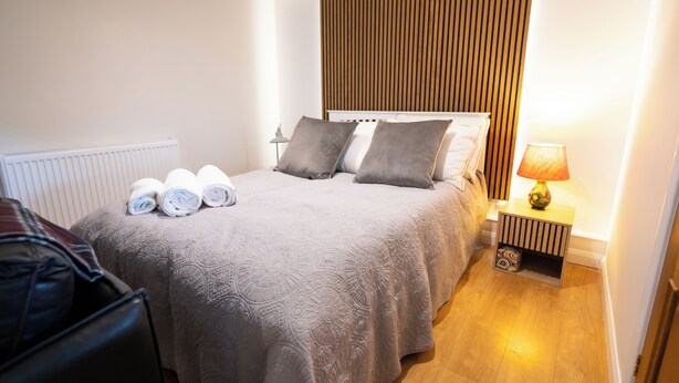 A neatly made bed with a grey quilt, two pillows, and rolled towels on the side. There is a bedside table with a lamp and a small decorative item. - Aethelred Annexe - New Build with Full Amenities (Winchester)