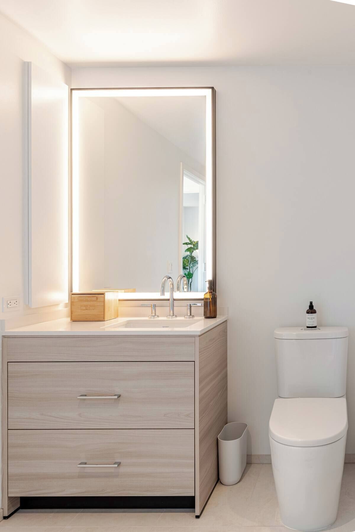 A modern bathroom with a wooden vanity, a large mirror, a white toilet, and a trash can.