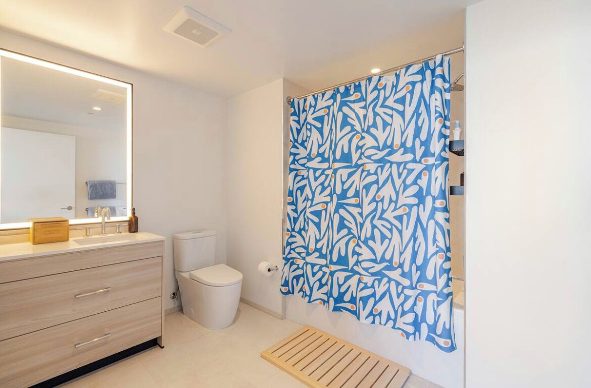 A modern bathroom with a large mirror, a wooden vanity, a toilet, and a shower with a blue leaf-patterned curtain.