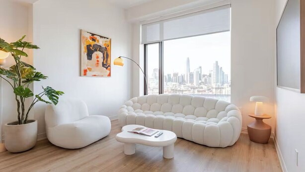 A modern living room with a white sofa, a white armchair, a coffee table, and a large window with a city view.