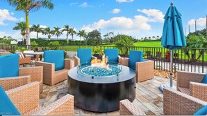 An outdoor patio with wicker furniture, a central fire feature, and a view of a green field and palm trees. - Countryside Golf and Country Club 2bed 2bath all new Clubhouse & facilities (Naples)