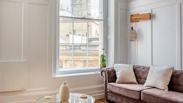 Een kamer met een groot raam, een bruine bank met witte kussens, een glazen salontafel en een hanglamp.