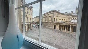 Uitzicht door een raam met een vaas op de vensterbank, op een kinderkopjesstraat met historische gebouwen.