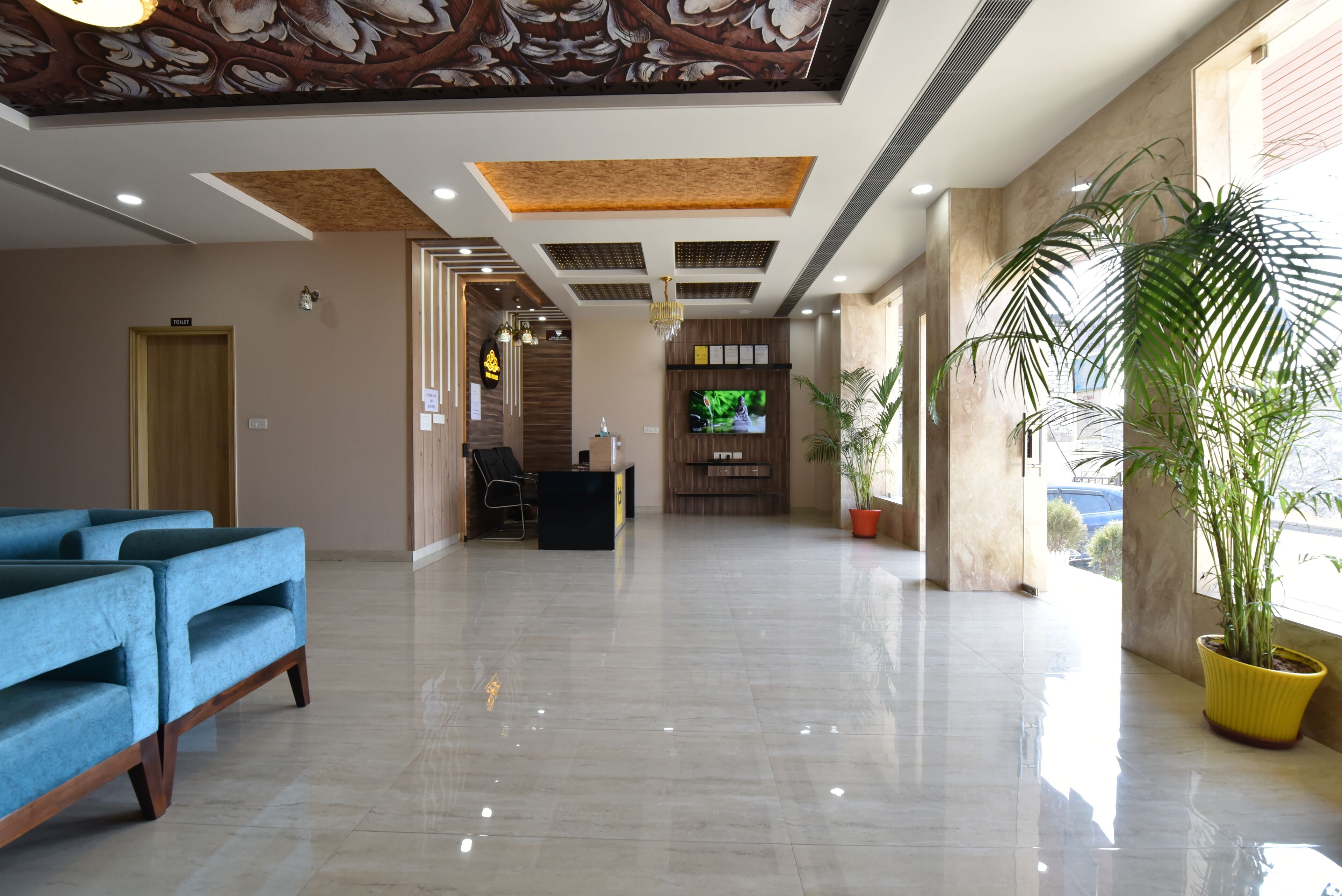 A spacious lobby with a reception desk, blue sofas, a large potted plant, and a TV screen.