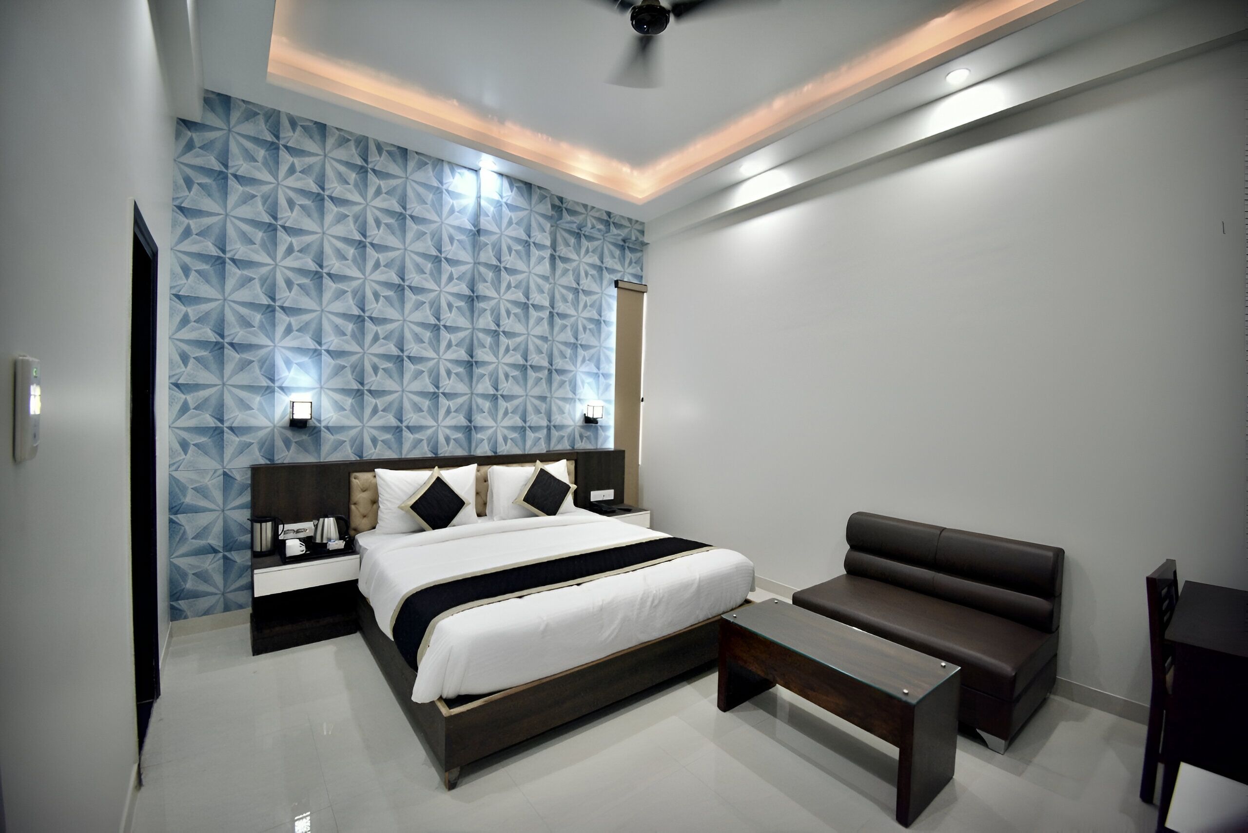 A modern bedroom with a geometric-patterned wall, a large bed, a dark brown sofa, and a small table.
