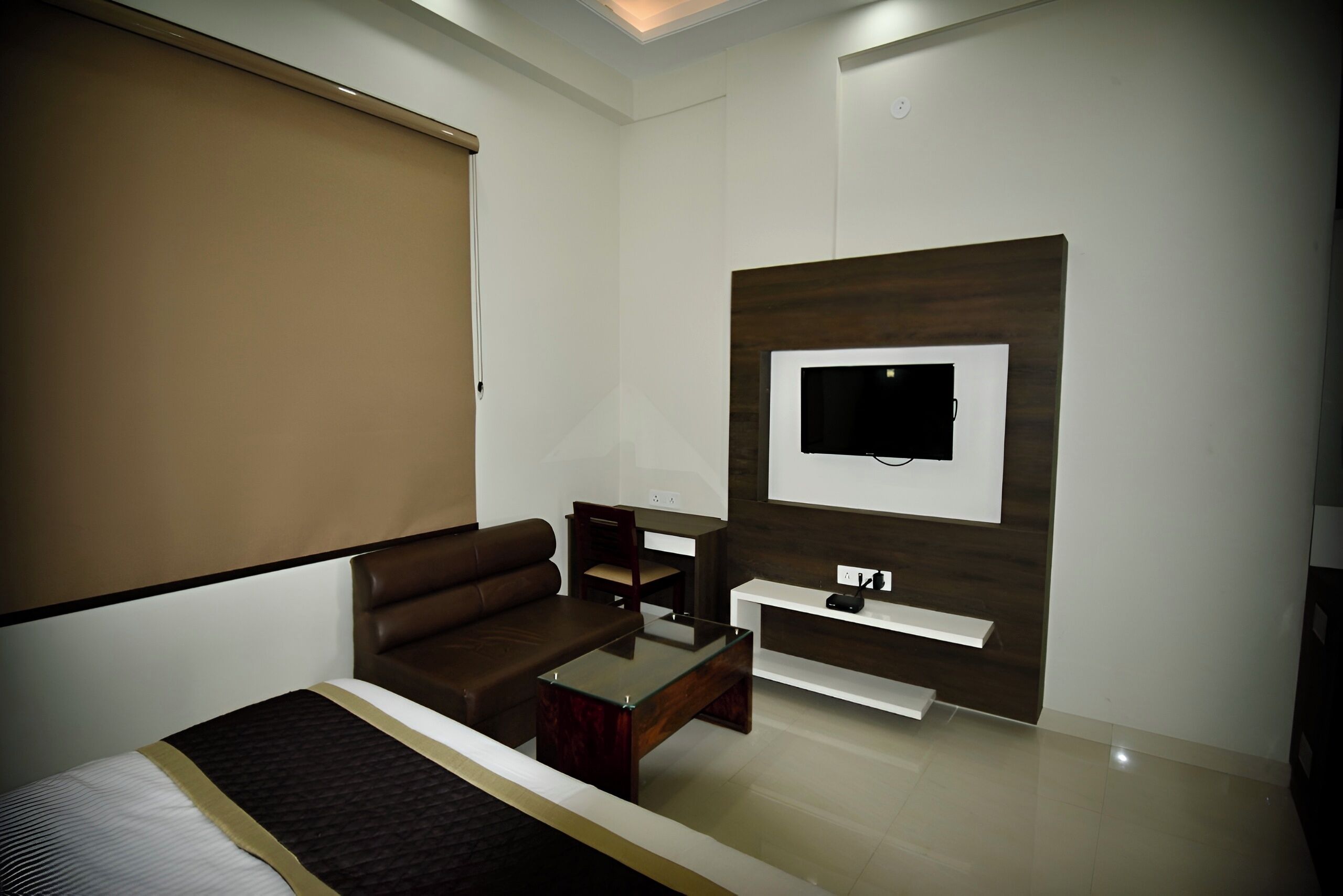 A modern hotel room with a bed, a brown leather sofa, a glass coffee table, a TV mounted on a wooden wall, and a white console table.