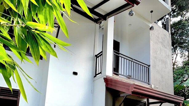 Une maison à deux étages avec un balcon, un petit patio avec une table et des chaises, et un jardin agrémenté de plantes en pot.