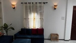 Un salon spacieux avec un grand canapé d’angle, un ventilateur de plafond et une grande fenêtre agrémentée de rideaux légers.