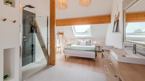 Une chambre moderne dotée d’une douche en verre, de poutres en bois, d’un puits de lumière, d’un grand lit, d’une table de chevet et d’une salle de bain avec un grand miroir et un lavabo.
