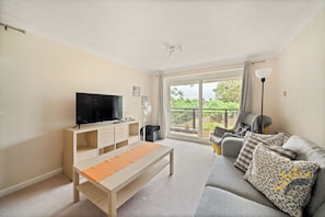 A living room with a TV, sofa, and a balcony with a view of greenery.