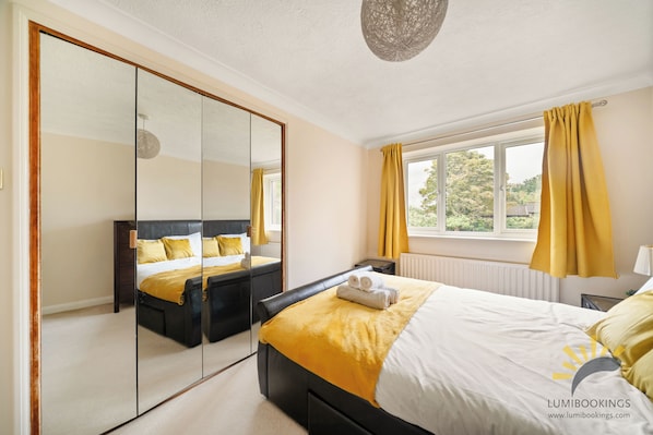 A modern bedroom with a large mirror, a bed with yellow bedding, a window with curtains, and a radiator.