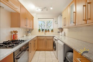 A modern kitchen with wooden cabinets, a gas stove, a washing machine, and a window with plants.
